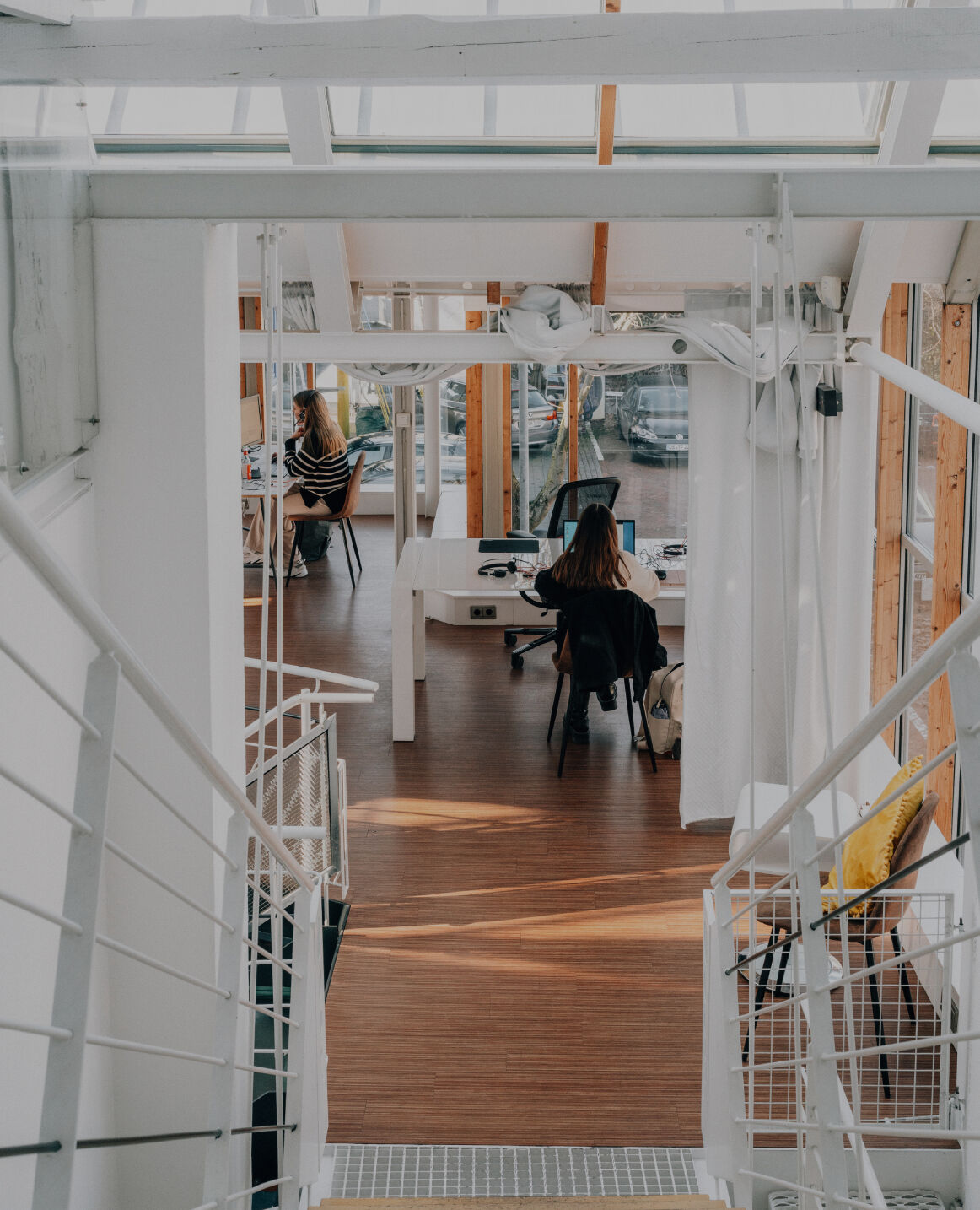 Blick von einer Treppe in ein helles, modernes Büro mit Holzfußboden, Glasfronten und zwei Personen an Schreibtischen