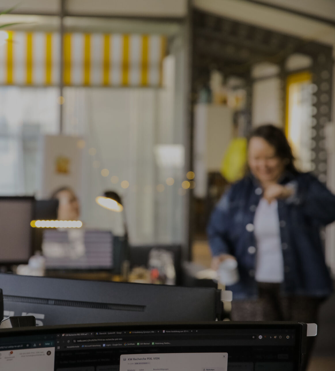 Büroszene mit Monitor im Vordergrund und einer Person, die mit einer Tasse durch das Büro geht