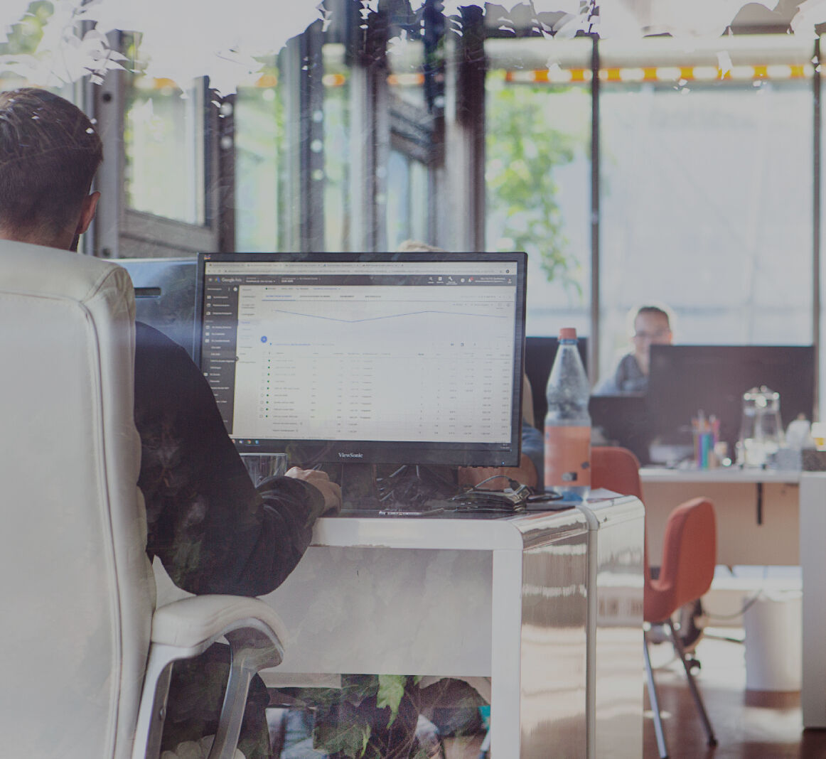 Heller Büroraum mit mehreren Arbeitsplätzen, im Vordergrund eine Person am Schreibtisch vor einem Monitor mit Tabellenansicht