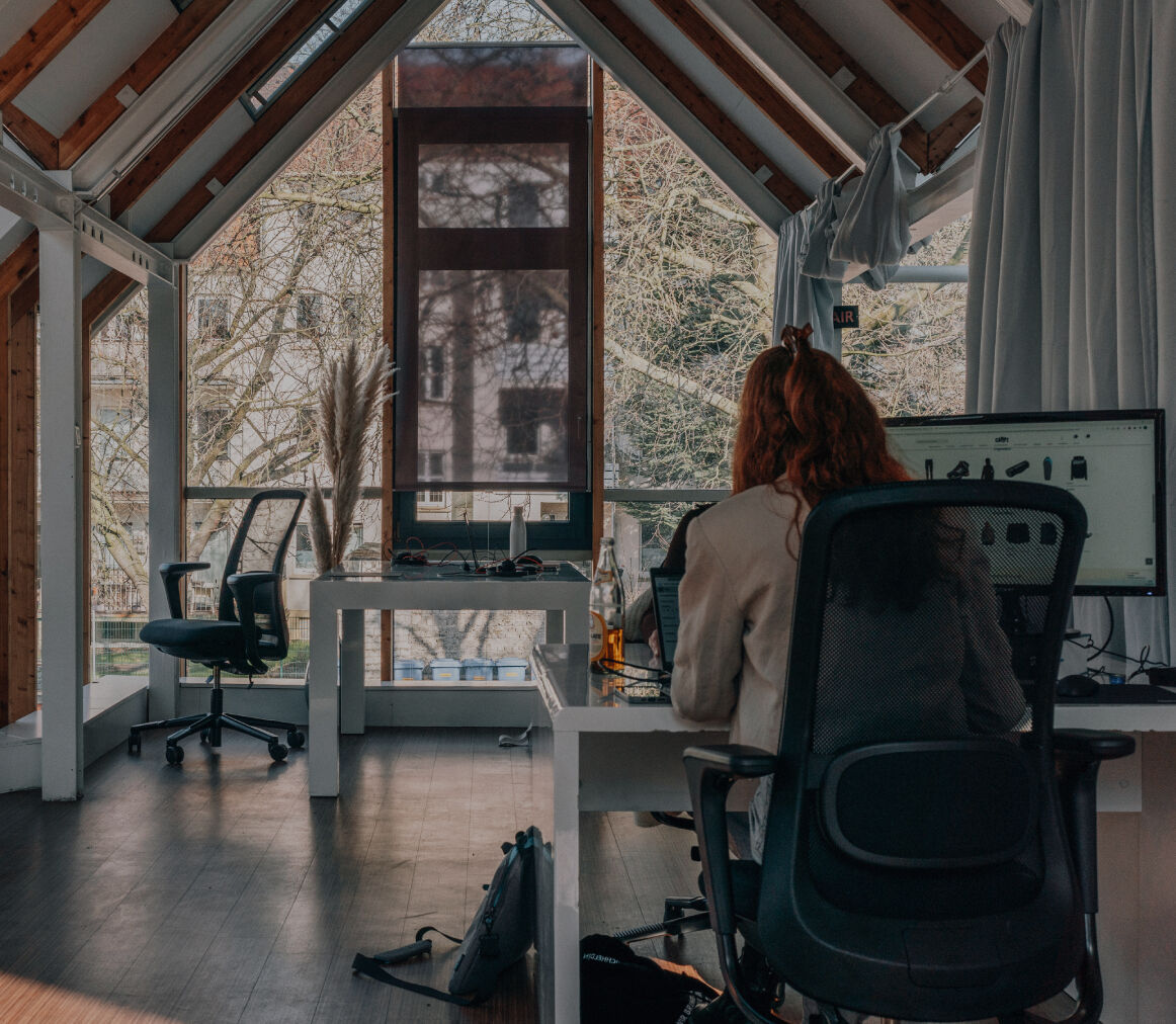 Person arbeitet an einem Schreibtisch im Büro, seitlich durch eine Glaswand fotografiert, mit Fensterfront im Hintergrund.