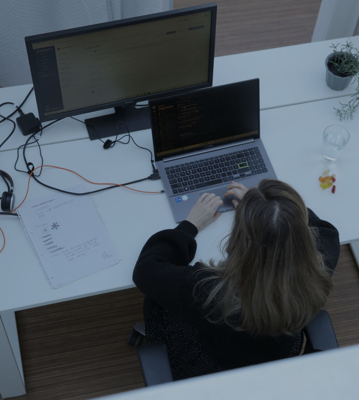 Person tippt an einem Laptop am Schreibtisch im Büro, aufgenommen aus der Vogelperspektive, mit externem Monitor und kleiner Pflanze