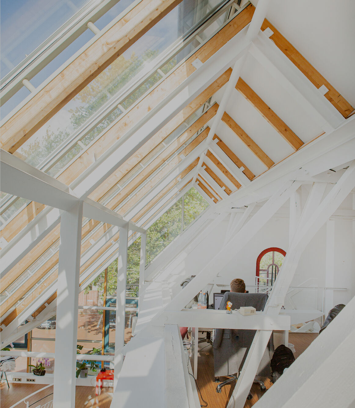 Helles Dachgeschossbüro mit großen Glasdachfenstern und sichtbaren Holz- und Stahlträgern; eine Person sitzt an einem Schreibtisch, viel Tageslicht fällt in den offenen, modernen Arbeitsbereich